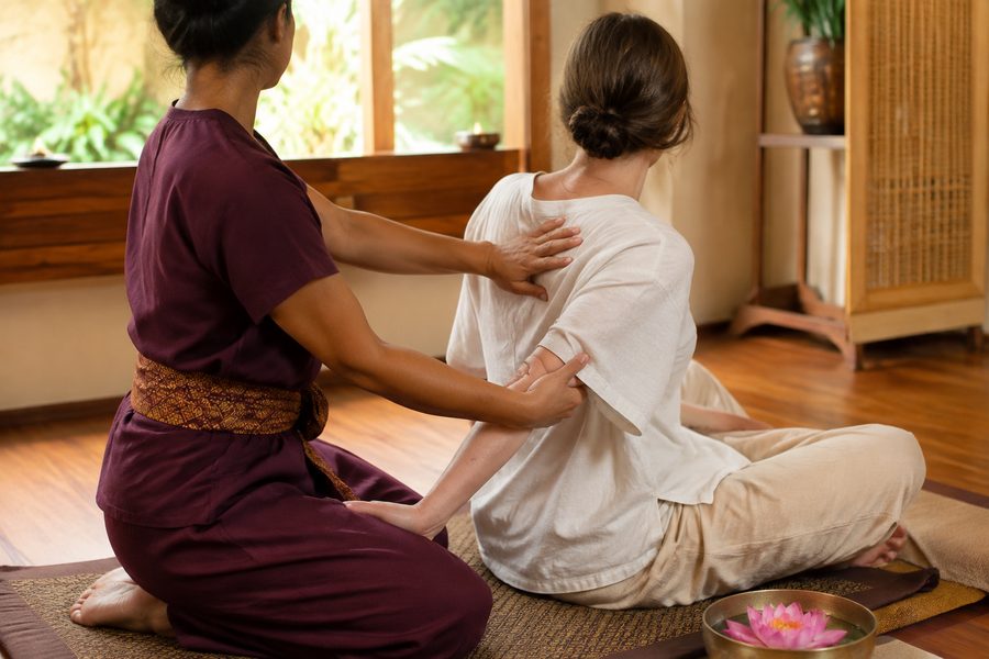 Therapist guiding a client through a seated Thai massage stretch on a floor mat